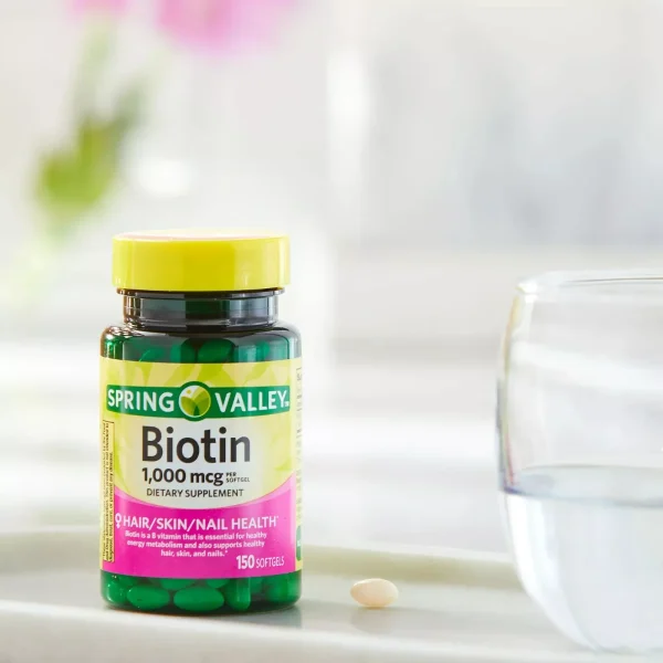 A bottle of Spring Valley Biotin 1000 Mcg softgels sits on a tray beside a glass of water, promoting healthy skin, hair, and nails.